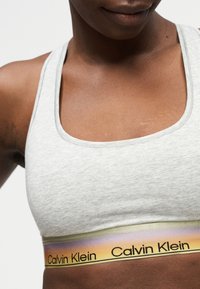 Κοντινό πλάνο ενός ατόμου που φοράει αθλητικό σουτιέν σε ανοιχτό γκρι χρώμα, με ελαστική ταινία κάτω από το στήθος, με το λογότυπο Calvin Klein σε αποχρώσεις διαβαθμισμένα χρώματα.