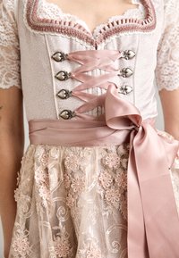 Jasnoróżowy dirndl z metalowymi akcentami w kształcie serca, koronkowym wykończeniem i satynowym paskiem. Zawiera kwiatowe hafty na przezroczystej spódnicy.