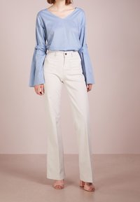 Haut bleu clair à manches longues avec des manches évasées, associé à un pantalon blanc taille haute. Le mannequin porte des sandales à talons blocs rose clair.