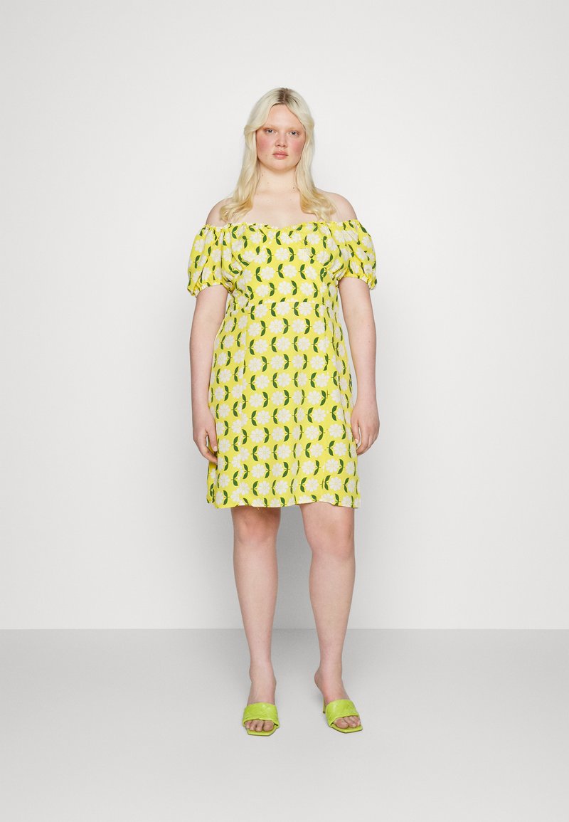 Vestido de ombros à mostra em amarelo com padrão floral branco e detalhes verdes; comprimento curto e mangas bufantes, combinado com sandálias verdes de slide.