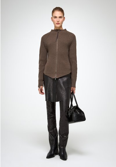 Maglione zippato a coste marrone con colletto alto, abbinato a una gonna in pelle nera e pantaloni in pelle nera aderenti, e una borsa nera.