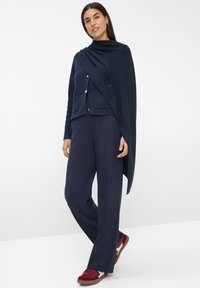 Marineblaues Strickcardigan mit tailliertem Schnitt, goldenen Knöpfen und einer Tasche. Kombiniert mit weit geschnittenen marineblauen Hosen und burgunderfarbenen Sneakers.