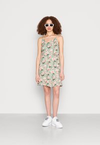 Vestido floral verde com alças finas e bainha em babados, combinado com ténis brancos. Óculos de sol estão a ser usados. Tecido texturizado com padrões vibrantes.
