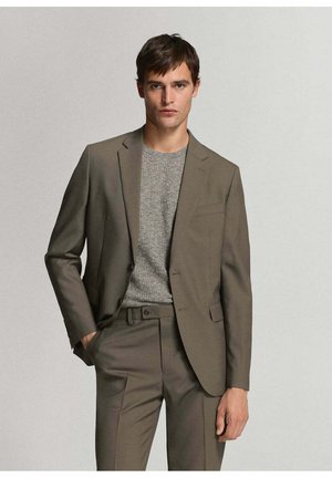 Joven con traje verde oliva y suéter de punto gris, de pie con una mano en el bolsillo frente a un fondo liso y claro.