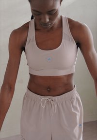Sujeción deportiva en beige pálido con paneles de malla, textura suave y un logo redondo en azul en el centro. Emparejado con pantalones cortos beige a juego.