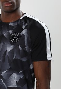 Czarny sportowy t-shirt z teksturowanym geometrycznym wzorem, z krótkimi rękawami i białymi akcentami. Na piersi znajduje się okrągłe logo.