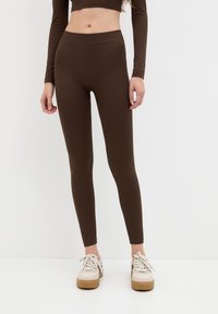 Nainen, jolla on yllä korkeavyötäröiset ruskeat legginsit ja niihin sopiva pitkähihainen crop-top, valkoiset lenkkarit jalassa, seisoo yksinkertaista valkoista taustaa vasten.