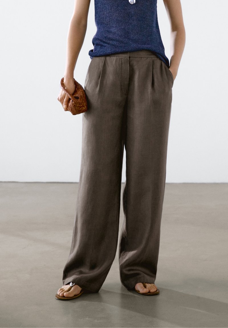 Grijze linnen broek met wijde pijpen, plooien en een hoge taille, gecombineerd met bruine teenslippers en een gestructureerde bruine handtas. Het bovenstuk is van blauwe tricot.