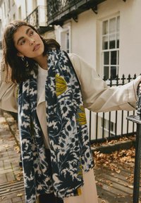 Uldscarf med en marineblå baggrund og gule blomster mønstre samt frynsekanter, båret med en beige frakke, sat mod en udendørs baggrund.