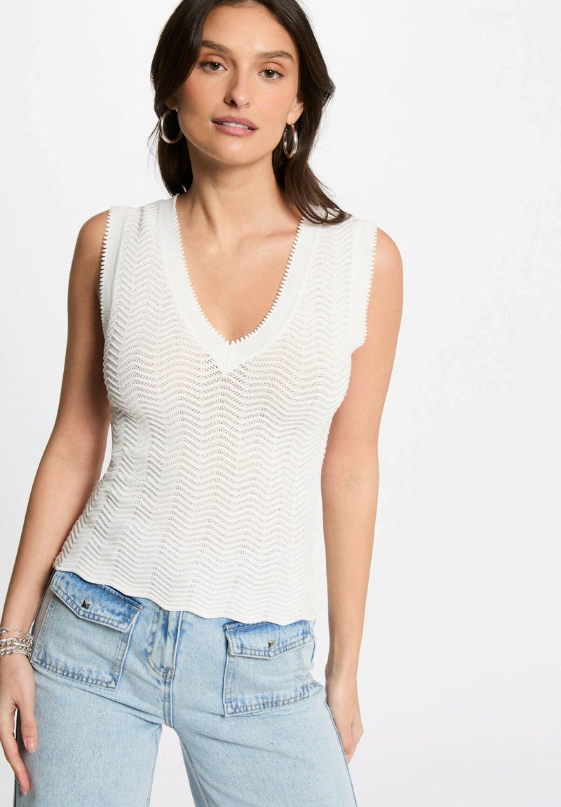 Top bianco senza maniche, con scollatura a V e un motivo a zigzag testurizzato, abbinato a jeans azzurri chiari a vita alta con tasche a toppa.