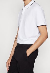 Vit polo-skjorta med svart krage och ärmsnibbar, med en plissering med två knappar. Tyget verkar slätt med en något strukturerad yta.