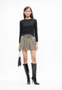 Egy nő fekete hosszú ujjú felsőt, kockás redőzött miniszoknyát övvel, fekete magassarkú térdig érő csizmát visel, és egy kis táskát tart a kezében.