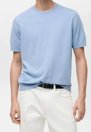 Man draagt een lichtblauw gebreid shirt met korte mouwen, lichtelijk ingestopt in een witte broek met een zwarte riem, staand tegen een effen achtergrond.