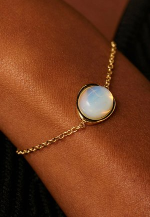 Bracelet en or avec une pierre ronde opalescente sertie dans une monture lisse semblable à un bezel, attaché à une chaîne délicate.