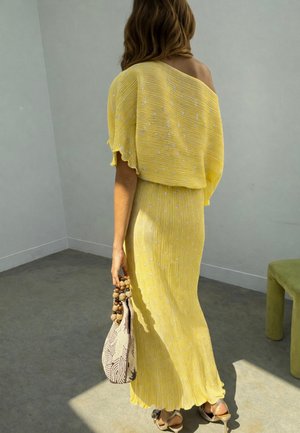 Femme en robe jaune épaules dénudées avec des pois blancs, tenant un sac beige avec des perles en bois, debout dans une pièce minimaliste avec un sol gris.