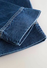 Jean en denim bleu plié montrant les coutures détaillées et la texture du tissu sur une surface claire.