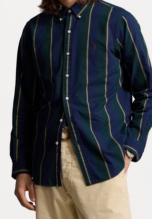Mężczyzna ubrany w granatowo-zieloną koszulę w paski zapinaną na guziki, beżowe sztruksowe spodnie oraz zegarek na lewej ręce.