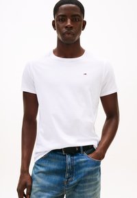 Vit t-shirt med rund halsringning i bomull, med en liten färgad logotyp på bröstet. Matchad med blåa jeans och ett svart bälte.