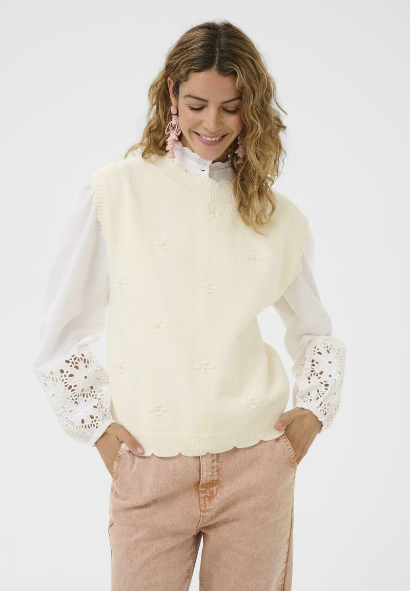 Vaalean kermanvärinen villapaita, jossa on kukka-ommeltuja yksityiskohtia, ribbireunus ja aaltoileva helma, päällä valkoinen pitsireunuksinen pusero ja vaaleanpunaiset housut.