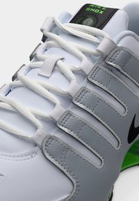 Λευκό και γκρι αθλητικό παπούτσι Nike Shox με μαύρο σήμα Swoosh, λευκά κορδόνια και πράσινες λεπτομέρειες σήμανσης στη γλώσσα και στη σόλα.