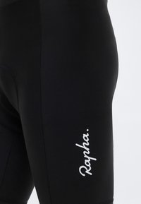 Κοντινό πλάνο μαύρου ποδηλατικού σορτς με το λευκό λογότυπο "Rapha." τυπωμένο κάθετα στον μηρό, πάνω σε ανοιχτό φόντο.