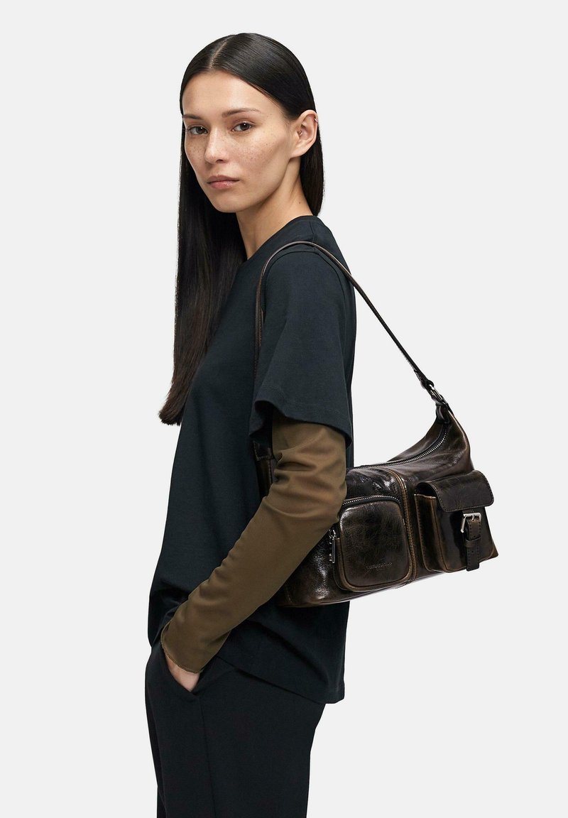 Czarna skórzana torba na ramię z wieloma kieszeniami, regulowanym paskiem i metalowymi dodatkami. Modelka ma na sobie czarną koszulkę z krótkim rękawem na brązowych długich rękawach.