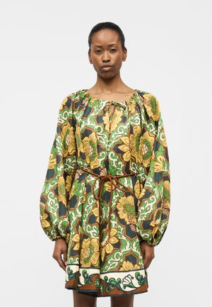 Femme portant une robe ample à motif floral avec des tons verts, jaunes et marron, dotée de longues manches bouffantes et d'une ceinture nouée à la taille.