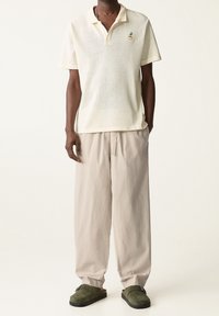 Kurzärmliges, hellbeiges Polohemd mit strukturiertem Mesh-Design und grüner Ananas-Stickerei, kombiniert mit lockeren beigen Hosen und grünen Schuhen.