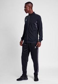 Schwarzer Trainingsanzug mit einer Jacke mit Reißverschluss und Hose, die an den Seiten hellgraue Zickzackakzente und ein Logo auf der Brust aufweist.