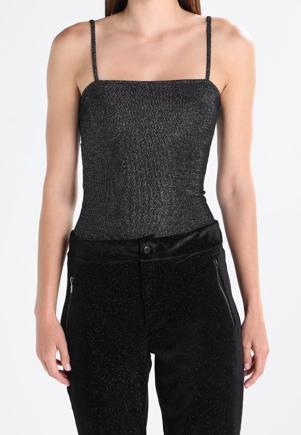 Femme portant un débardeur noir scintillant sans manches associé à un pantalon noir taille haute avec poches zippées.