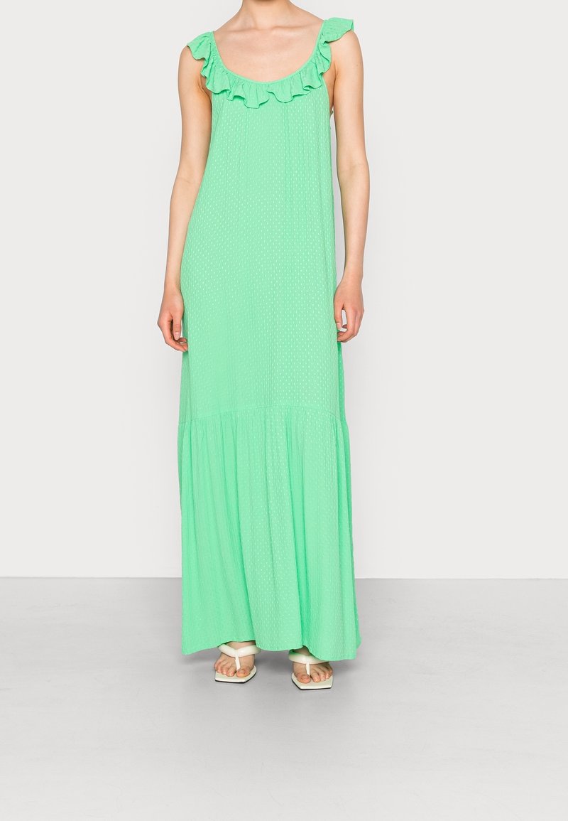 Femme portant une robe maxi sans manches vert clair avec un décolleté à volants et des sandales blanches à bout ouvert, debout sur un fond uni.