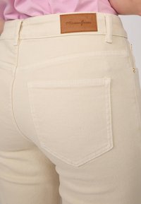 Pantalon en denim couleur crème avec une finition texturée, doté d'une étiquette en cuir marron et de deux poches arrière. Détails de couture soignés.