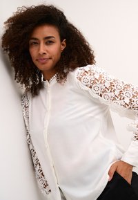 White blouse featuring lace sleeves with floral patterns, button front, and a smooth texture, paired with black pants.