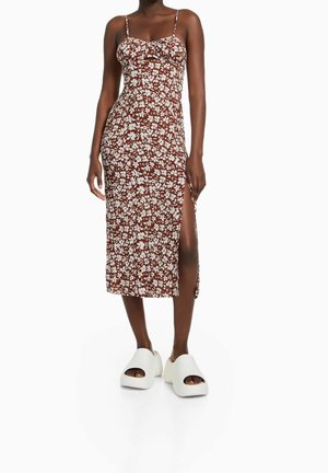 Mujer vestida con un vestido midi marrón con estampado floral, tirantes finos y abertura lateral, combinado con zapatos de plataforma blancos, gruesos y de puntera abierta.