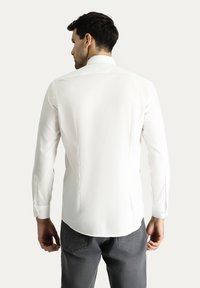 Camisa blanca de manga larga hecha de una tela suave con cuello puntiagudo, costuras en la parte posterior y puños abotonados. Combinada con pantalones grises.