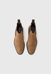 Botas Chelsea de ante marrón con paneles laterales elásticos y tiradores, que presentan una puntera redonda y detalles de costura a lo largo de la suela negra.
