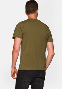 Oliwkowa koszulka z krótkim rękawem, wykonana z miękkiej bawełny, z okrągłym dekoltem i minimalistycznym wzornictwem, widoczna z tyłu, w połączeniu z czarnymi dżinsami.