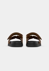 Brune lædersandaler med udskåret design, der har justerbare stropper og en sort gummisål. Set bagfra, hvilket viser struktur og form.
