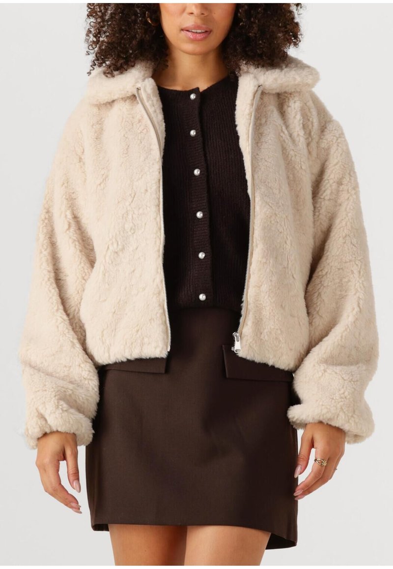 Crème-kleurige nepbontjas met een ritssluiting aan de voorkant, een gestructureerde kraag en oversized mouwen, gecombineerd met een zwarte blouse met knopen en een bruine rok.