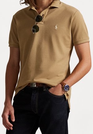Homme portant un polo beige à manches courtes avec logo, jean foncé, montre en cuir marron, ceinture marron et lunettes de soleil rondes accrochées à la chemise.