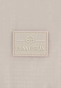 Vaalea kankaan tausta, jossa on suorakulmainen valkoinen logo-etiketti, johon on kohokuvioitu "JOHNNY URBAN" ja silmälasikuvio.