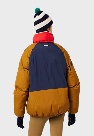 Veste matelassée composée de panneaux orange, bleu marine et rouge ; poignets côtelés ; avec un bonnet rayé en tricot orné d'un pompon vert.