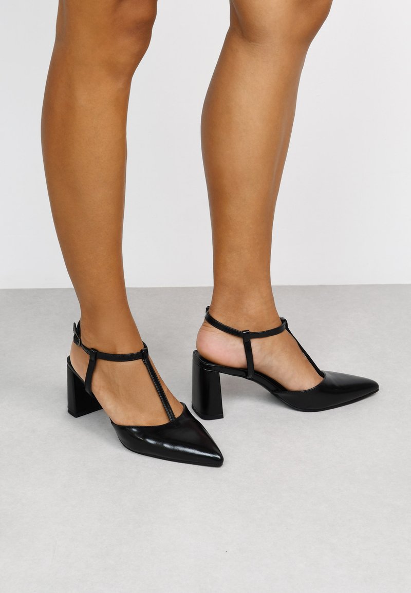 Zapatos de tacón de piel negra con punta afilada, diseño de tira en T y tacón ancho. Acabado brillante y correa ajustable en el tobillo.