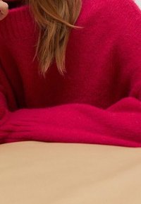 Maglione magenta a maglia con vestibilità ampia e polsini a coste, poggiato su una superficie beige chiaro. Texture morbida con fibre visibili.