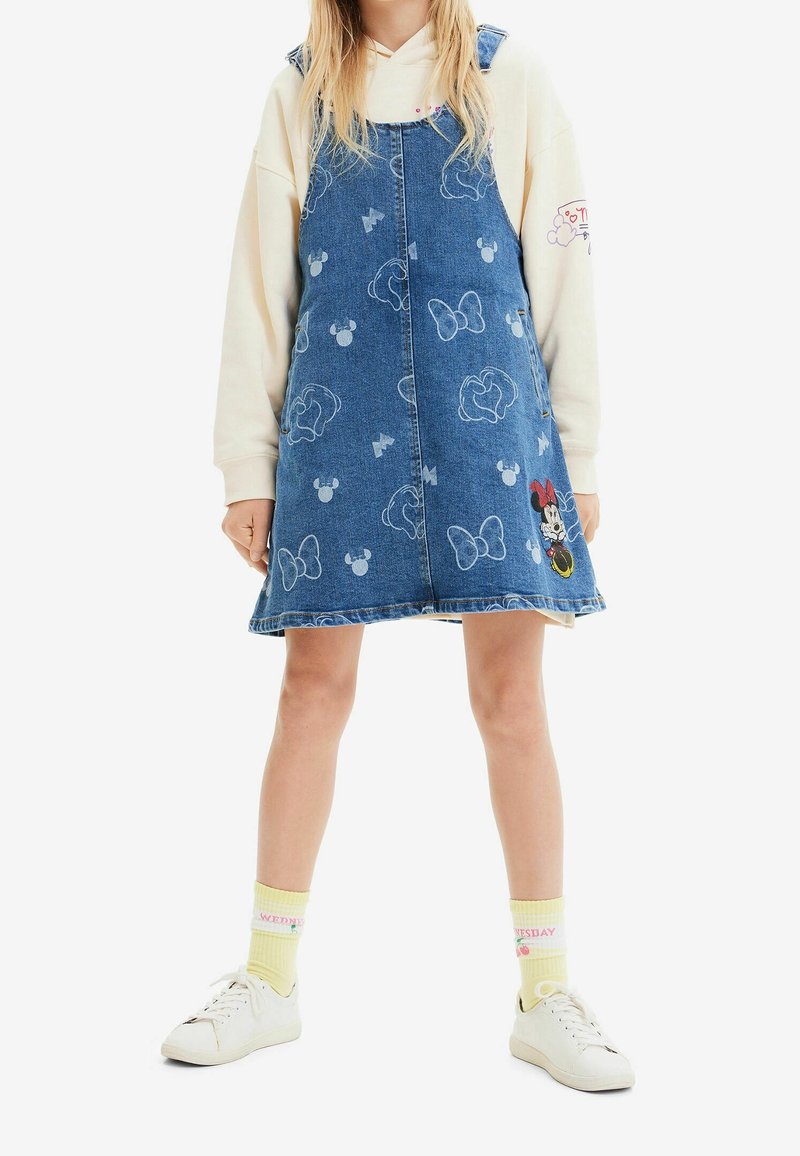 Vestido peto de mezclilla de color azul claro, con patrones de Mickey Mouse y lazo en blanco, cortado por encima de las rodillas, usado sobre una sudadera color crema.