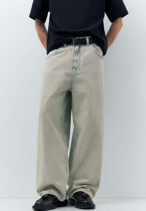 Persoon gekleed in lichtbeige wijde jeans, zwarte riem, zwart T-shirt met korte mouwen en zwarte leren schoenen met kwastjes, handen achter de rug.