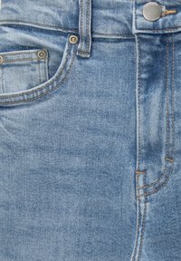 Jean en denim bleu clair en gros plan montrant la poche avant, la boucle de ceinture, le bouton et les détails de couture orange.