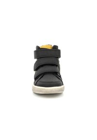 Zapatilla alta negra con tres correas de velcro, suela de goma y acentos amarillos. Parte superior de cuero texturizado y área de puntera perforada.