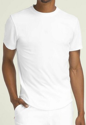 Mann trägt ein schlichtes weißes Kurzarm-T-Shirt mit der Hand in der Tasche vor hellem Hintergrund, Torso und Arme sichtbar.