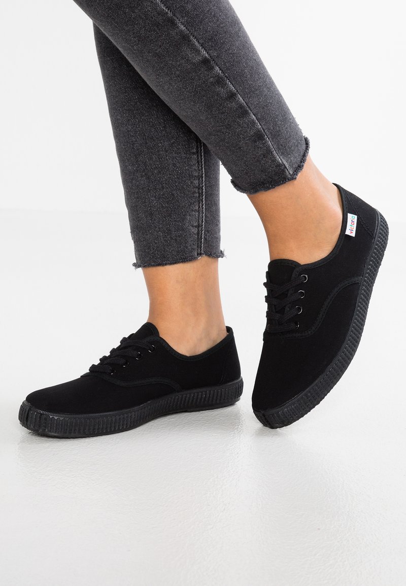 Svarta kanvas sneakers med rund tå, snörning framtill och texturerad gummisula. Har en liten färgtagg på sidan och en avslappnad design.